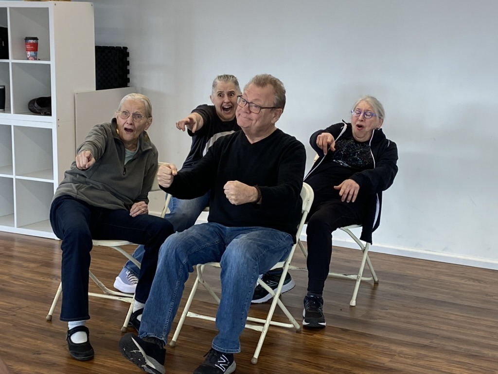 Four older adults sit in chairs. One man looks like he's driving with a bemused look on his face. The other three appear to be his passengers, and they're all screaming and pointing.